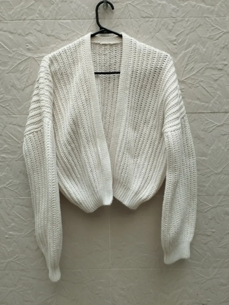 Cardigan a crochet de color marfil colgado de un gancho negro en una pared de color beige claro.