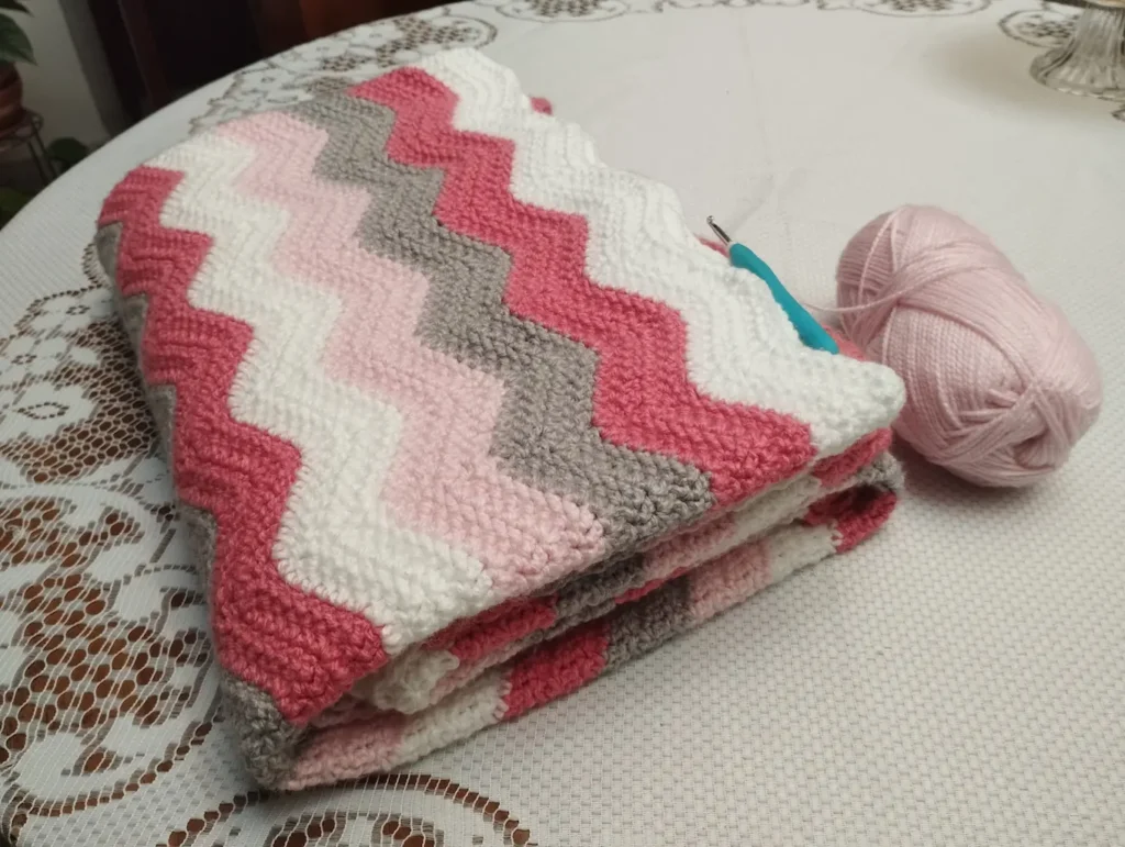 En el lado izquierdo hay una cobija a crochet de franjas en zig zag de colores: blanco, salmón, gris y rosado claro. La cobija está sobre una superficie blanca y en el lado derecho hay hay un ovillo de color rosado y una aguja de crochet con mango azul.