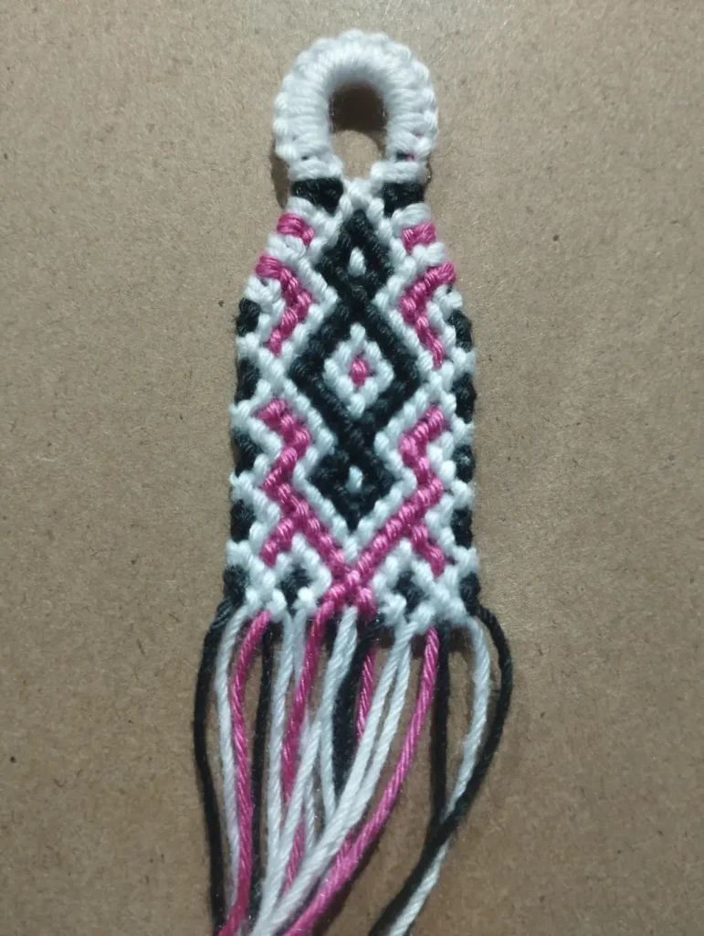 Tejido en macramé sobre una superficie color cafe. En la parte superior, el tejido tiene un aro de hilo de color blanco. Inmediatamente abajo del hilo hay un tejido con formas de rombos de colores blanco, negro y rosados.