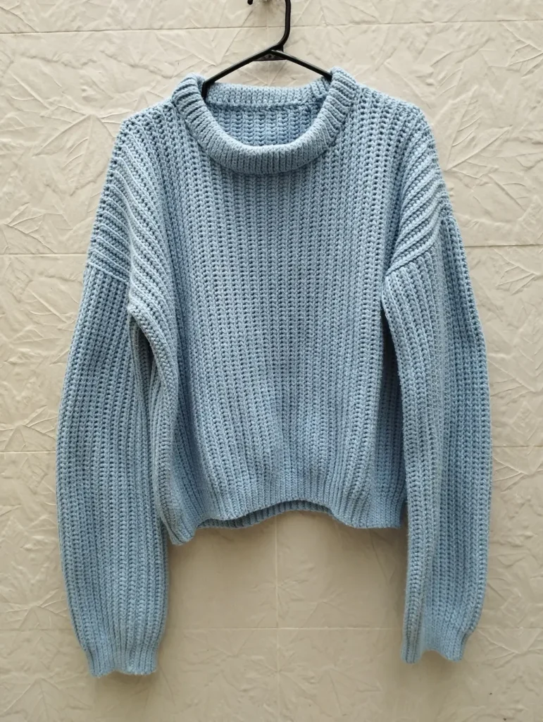 Sueter a crochet de color azul claro colgado de un gancho negro en una pared de color beige claro.