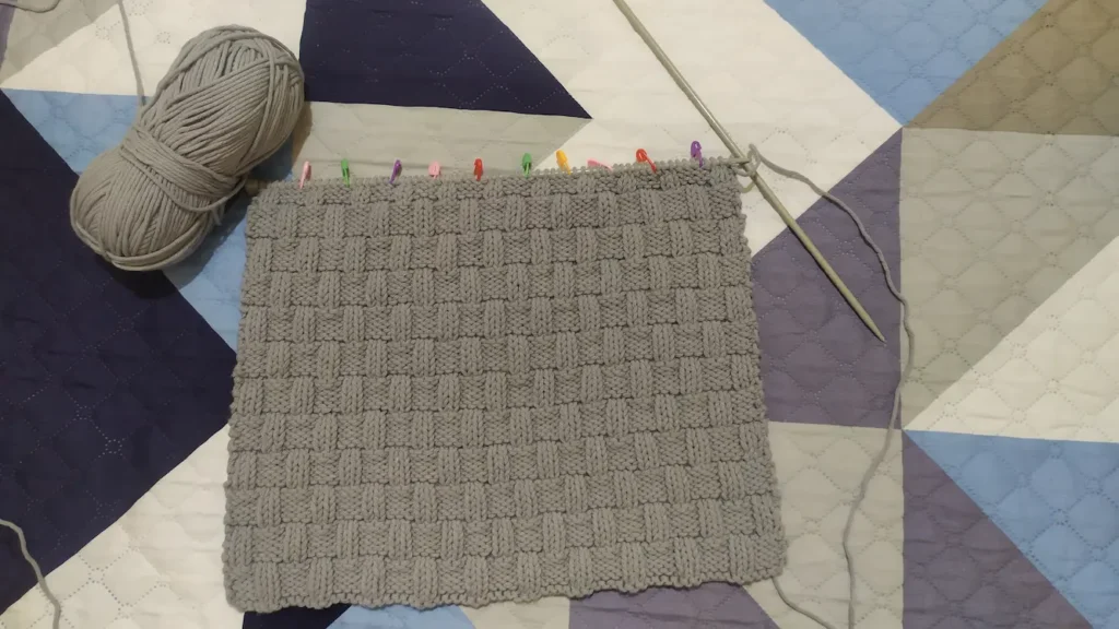 En el centro, tejido a dos agujas de color gris con textura de cesta. Cerca hay una aguja y un ovillo de color gris. El tejido está sobre un fondo geométrico de varias tonalidades de azul, gris y blanco.